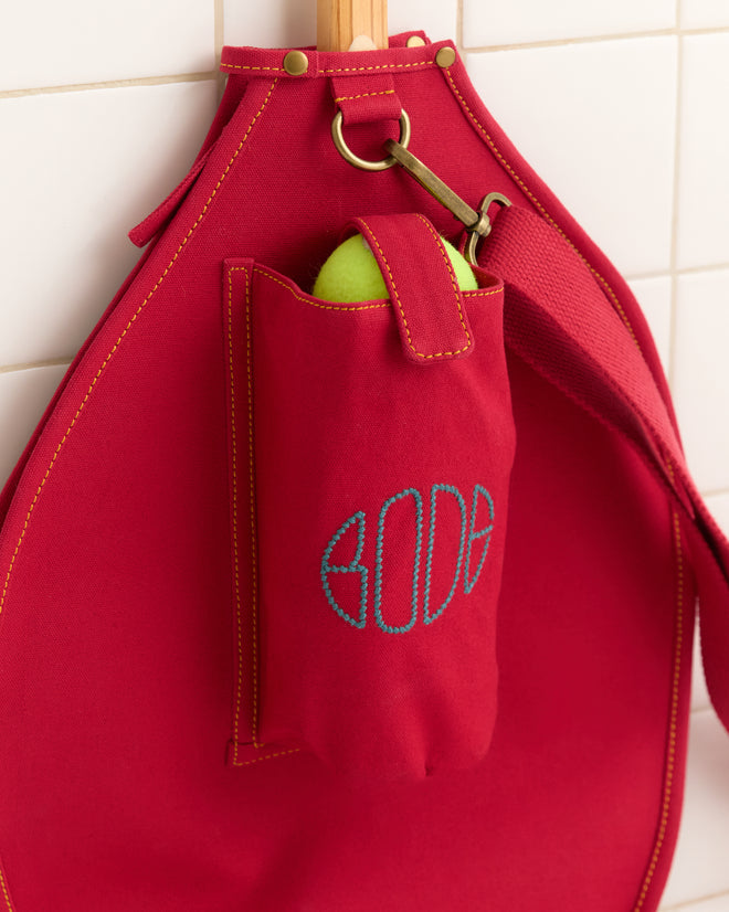 The Jacquard Racquet Sleeve, in red with an adjustable strap and pocket for a green tennis ball, hangs on a white tiled wall.