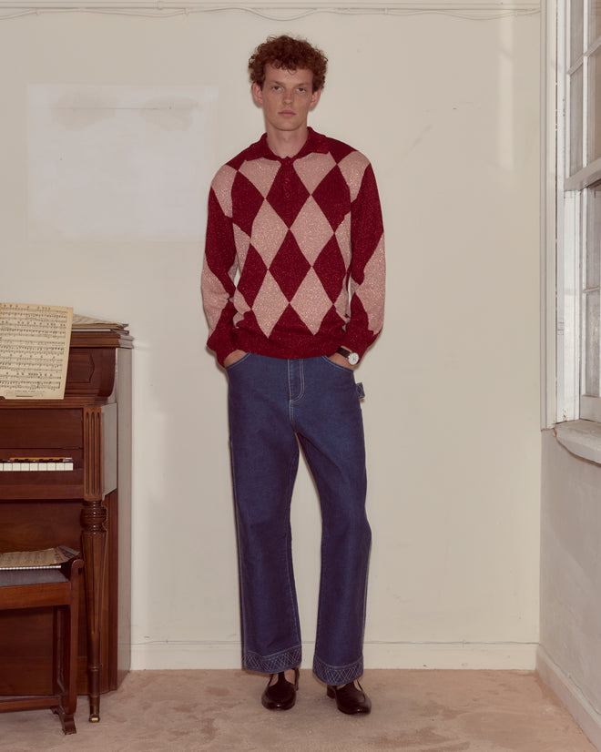 A man stands indoors by a piano, wearing a Jockey Polo, blue jeans, and brown shoes.