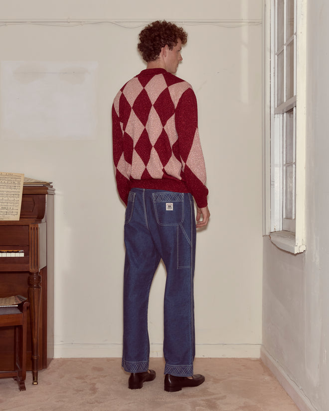 A person wearing a Jockey Polo with a red and white argyle pattern and blue jeans stands facing away in a sparsely furnished room.