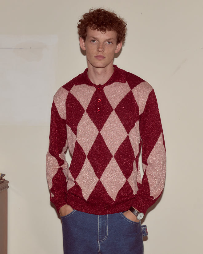 A person with curly hair wears a Jockey Polo and blue jeans, standing against a plain wall.