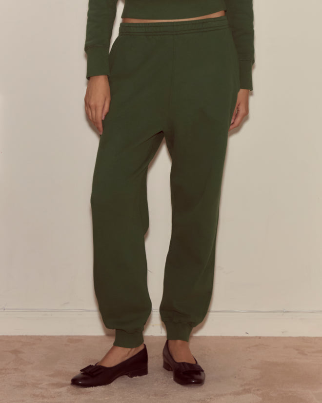 A person wearing Joggers - Dark Green and black flat shoes stands against a light wall on a carpeted floor.