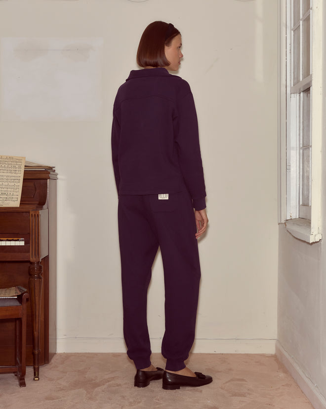 A woman in dark purple joggers and a navy tracksuit stands facing a wall next to a piano and window, on carpeted flooring.