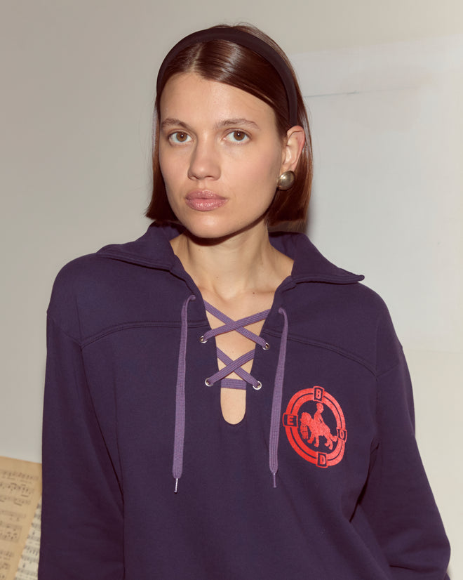A woman with straight brown hair in a headband wears a purple Pet Pullover featuring a lace-up collar and a red emblem, looking at the camera.