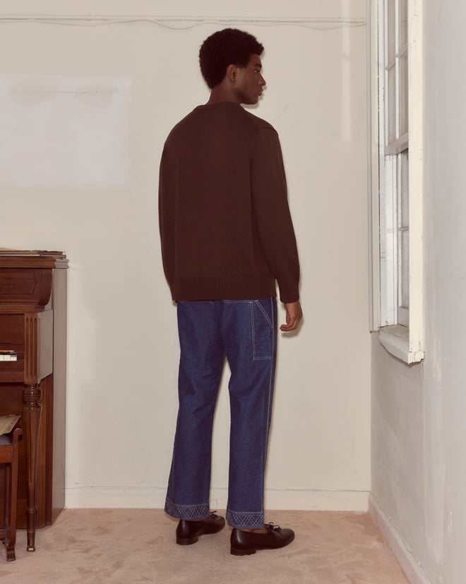 A man wearing the Juniper Sweater in Brown/Black and jeans stands indoors facing a window, his back to the camera.