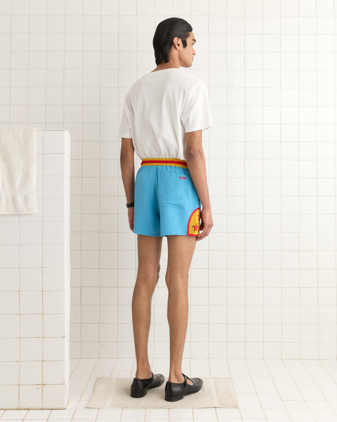 In a tiled room, an individual wearing a white shirt and Star Field Shorts - Blue, featuring appliquéd stars, stands facing away.