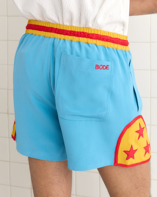 A person stands against a tiled wall, wearing Star Field Shorts - Blue, featuring yellow and red appliquéd stars.