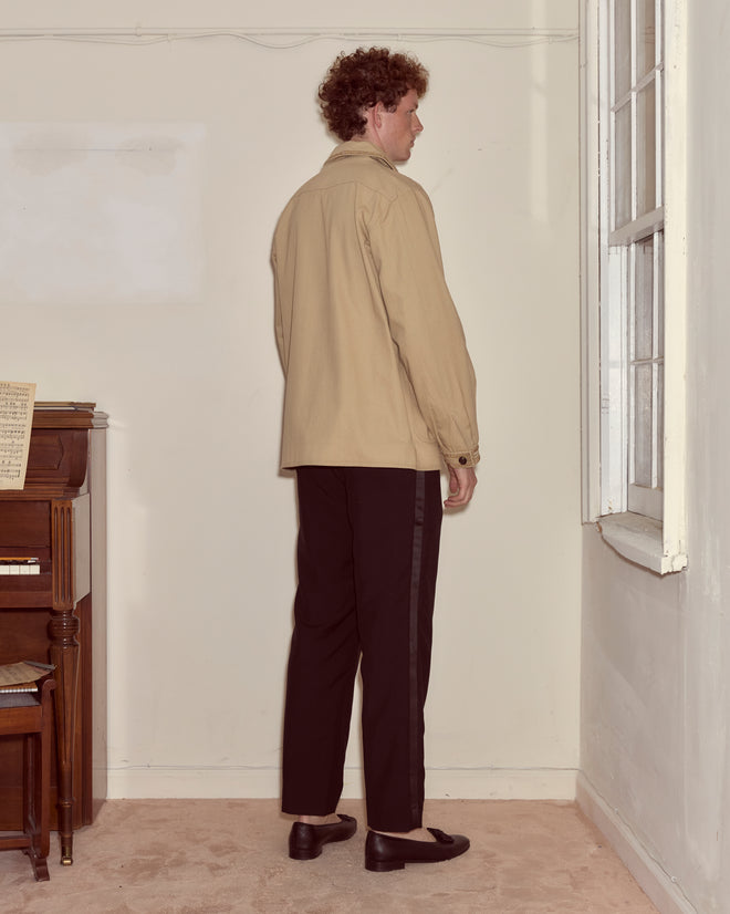 A man in a Khaki Bullion Jacket and black pants stands facing a wall in a room with a piano and large window.