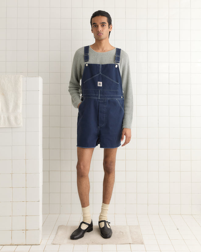 A person wearing Knolly Brook Overall Shorts stands on a pristine white tiled floor, their black shoes completing the look. In the background, a solitary towel hangs, adding a touch of casual charm to this workwear-inspired scene.