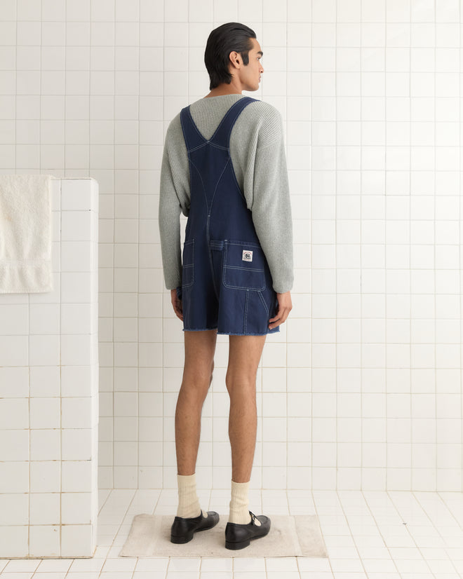 A person wearing a green merino wool sweater and Knolly Brook Overall Shorts stands in a tiled room, facing away.