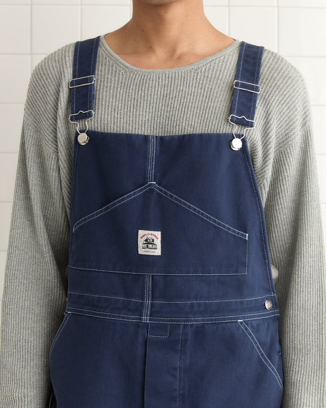 The person is clad in classic workwear, featuring a gray ribbed sweater and Knolly Brook Overall Shorts in blue denim with a pocket, standing against a tiled wall.
