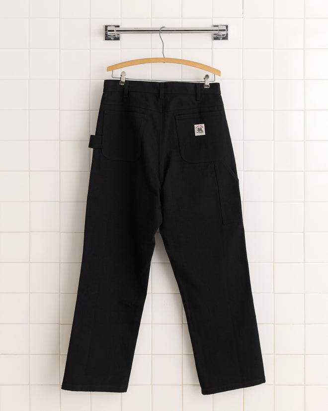 The Twill Knolly Brook Trousers - Black hang on a wooden hanger against a white tiled wall, their design evoking classic 1950s-1960s vintage style.