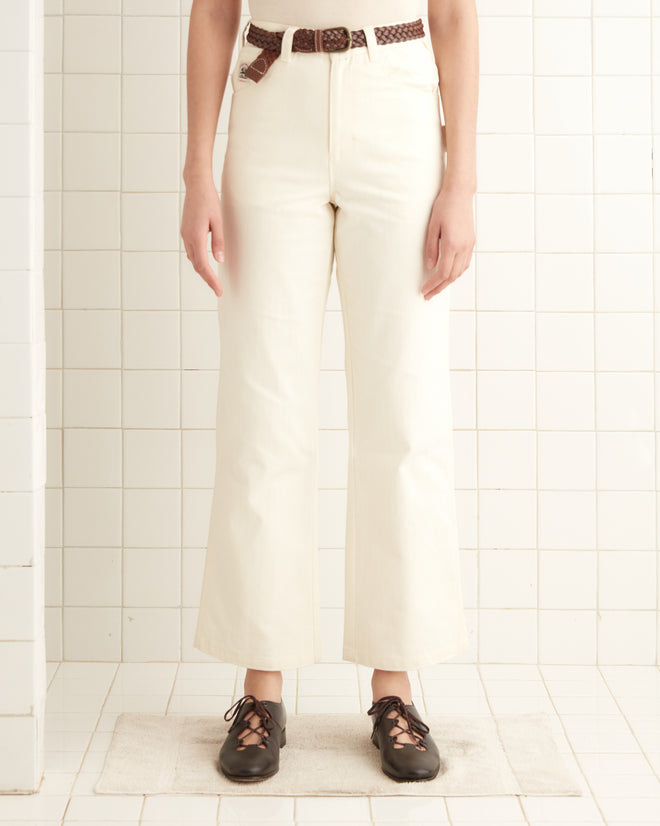 Dressed in Herringbone Knolly Brook Trousers - White, with a brown belt and shoes, a person stands on a small mat against a tiled wall.