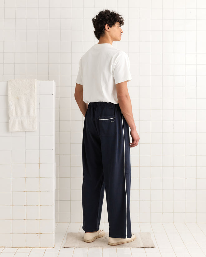 A person stands in a tiled room wearing a white shirt, white shoes, and navy Lace-Front Sweatpants with white stripes.