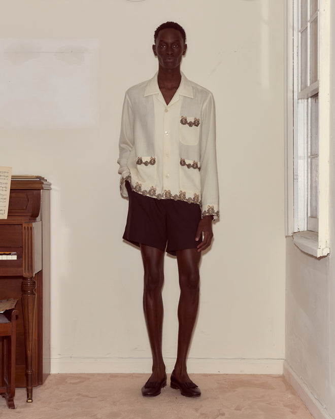 A person stands indoors by a piano, wearing the Laurelist Overshirt in cream with floral details and vintage passementerie, paired with black shorts and dark shoes.