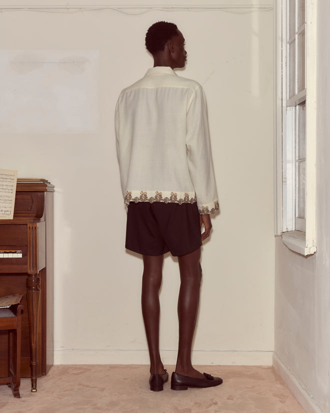 Wearing the Laurelist Overshirt in cream and black shorts, a person stands facing a wall beside a piano and a window adorned with vintage passementerie.