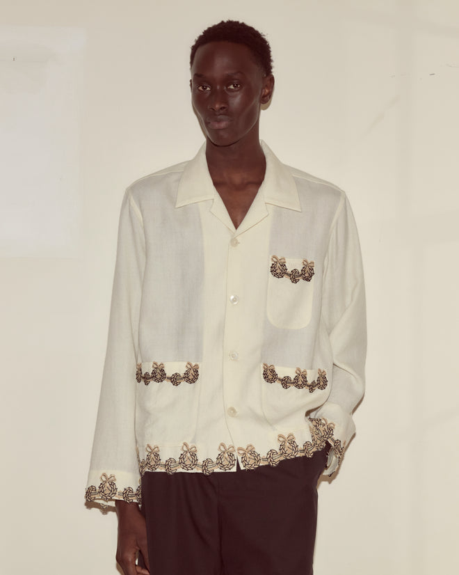 A person wears the Laurelist Overshirt in cream, featuring butterfly embroidery and vintage passementerie, paired with black pants against a light background.