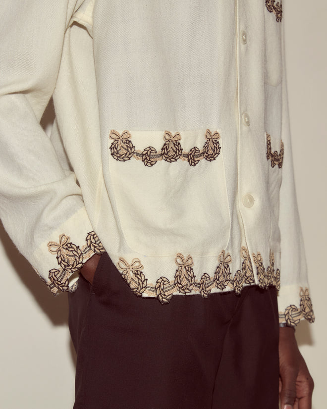 A person wears the Laurelist Overshirt in cream with brown embroidery and vintage passementerie, paired with dark pants, hand in pocket.
