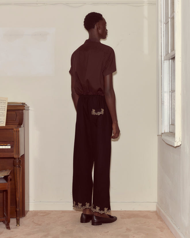 A person stands by a wall next to a piano, wearing Laurelist Pajama Pants in black, featuring gold embroidery at the waist and cuffs with vintage passementerie accents.