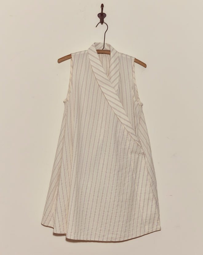 The Lavender Sail Dress, a sleeveless cream-colored striped wrap dress with antique pearl buttons, hangs on a wooden hanger against a plain wall.
