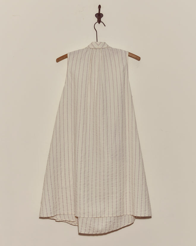 The Lavender Sail Dress is a sleeveless, high-neck, white 1930s-inspired dress with subtle vertical stripes and antique pearl buttons, displayed on a wooden hanger against the wall.