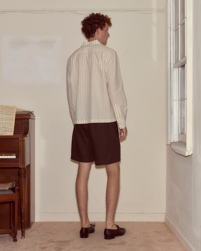 A man in a Lavender Sail Shirt and shorts stands facing the corner of a simple room near a piano and window.