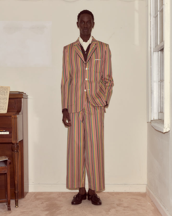 A person wearing the Lawrence Suit Jacket with colorful stripes and antique pearl buttons stands indoors by a piano and window, facing the camera.
