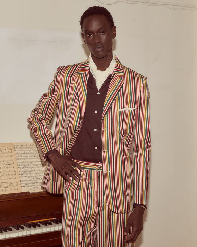 Wearing the Lawrence Suit Jacket, a person stands confidently by a piano with sheet music, showcasing the colorful single-breasted striped wool design and antique pearl buttons.