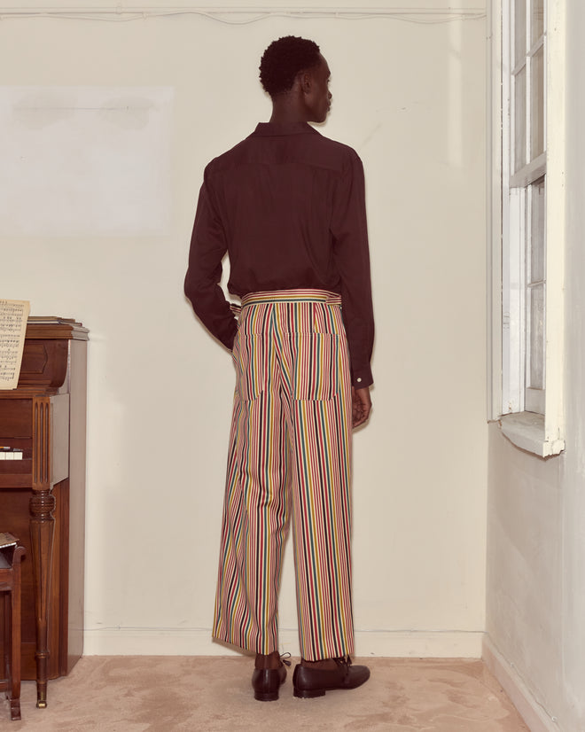 A person stands by a piano and window, facing a wall, wearing a dark shirt and the Lawrence Trousers—striped wool trousers with a wide-leg silhouette.