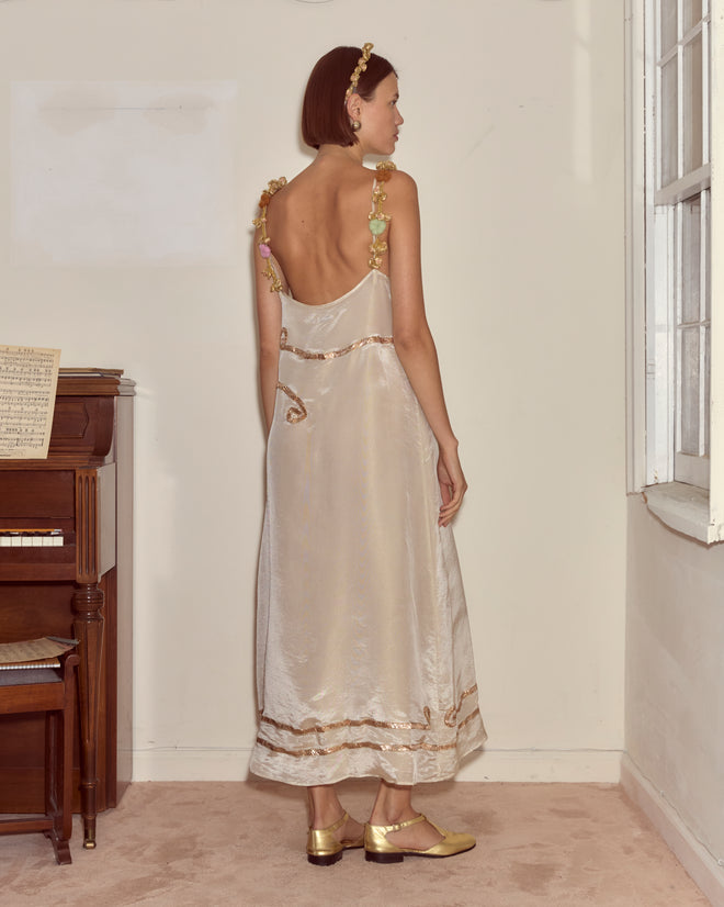 A woman in the Lido Dress, a cream appliqué gown with gold details, stands by a piano with her back to the camera.