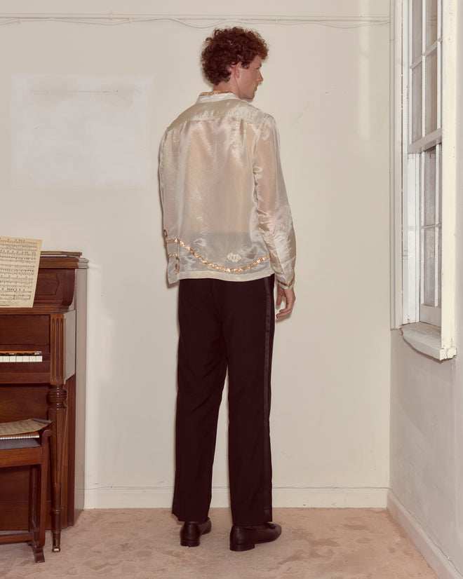 A man faces the corner of a beige room near a piano, wearing black pants and a shimmering Lido Long Sleeve shirt.