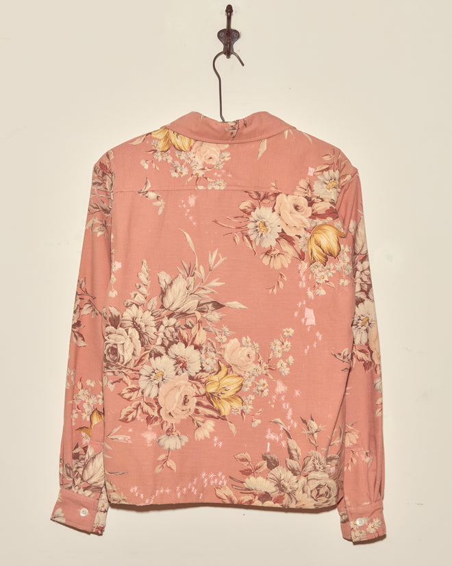 The Lily Rose Long Sleeve Shirt (XS/S), featuring a pink floral vintage tablecloth design, hangs on a wall hook and is displayed from the back.