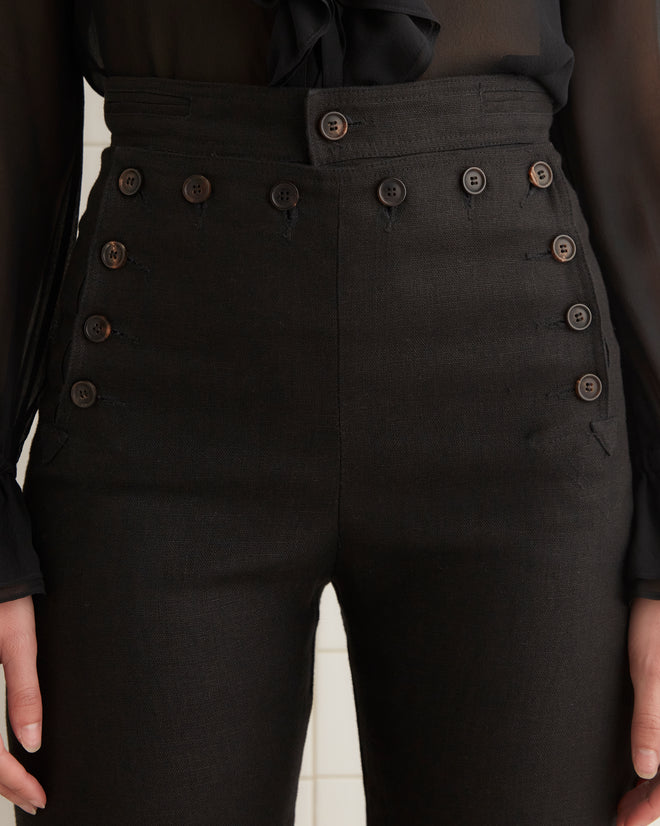 Someone is wearing Linen Sailor Trousers - Black with multiple buttons and a sheer black blouse, exuding style and confidence.
