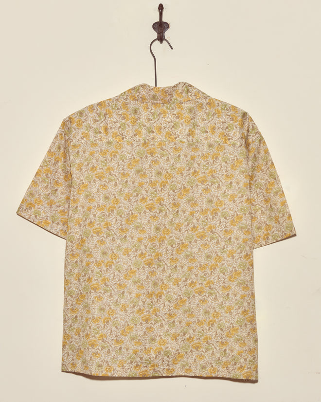 The Lion's Mane Floral Shirt (M/L), a short-sleeved vintage yellow floral piece, hangs on a hook against a plain light-colored wall.