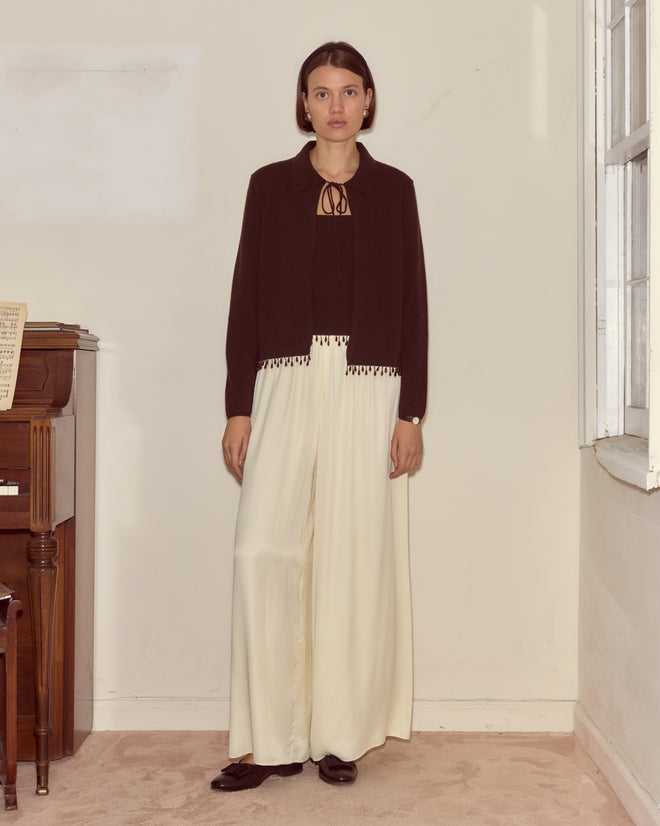 Indoors by a piano and window, a woman wears the Lodge Cardigan in dark brown.