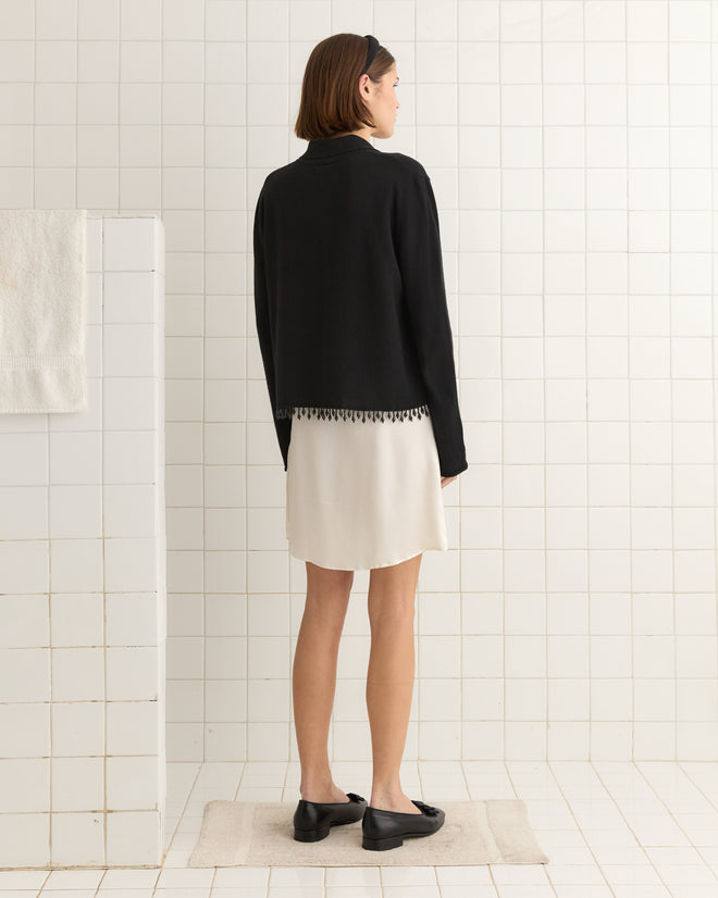 In a tiled room, someone is wearing a black Lodge Cardigan and white skirt, standing in front of a wall with a towel.