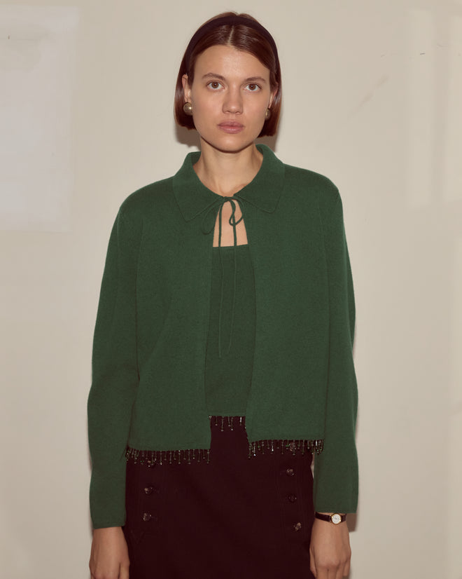 A woman with straight brown hair wears the Lodge Cardigan in dark green and a black skirt, standing against a plain light wall.