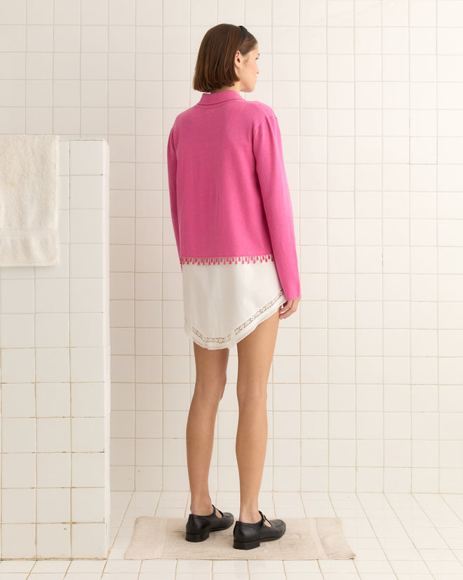 A woman in a Lodge Cardigan - Pink and a white dress stands on a bathroom mat, facing away in a tiled room.