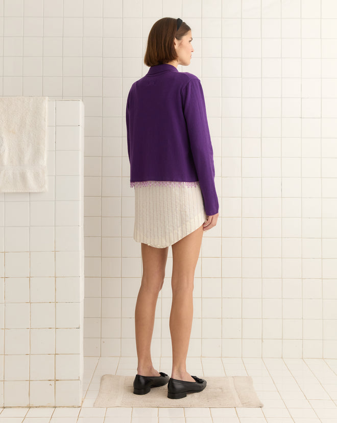 A woman in a Lodge Cardigan - Plum and a white skirt stands on a mat in a tiled room, facing away.