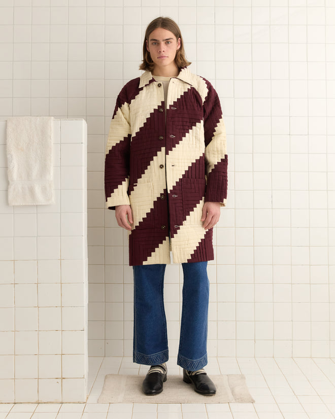 A person in a Log Cabin Suiting Coat and jeans stands in a tiled room with a towel on a ledge.