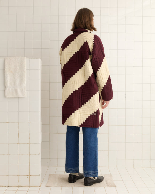In a tiled room, a person faces away wearing the Log Cabin Suiting Coat, which is maroon and beige with a quilted design and an embroidered logo.