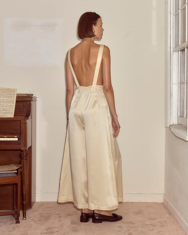A woman in the Loulou Jumpsuit - Cream stands indoors by a piano and window, facing away, showcasing the elegant open-back design inspired by refined 1960s jumpsuit style.