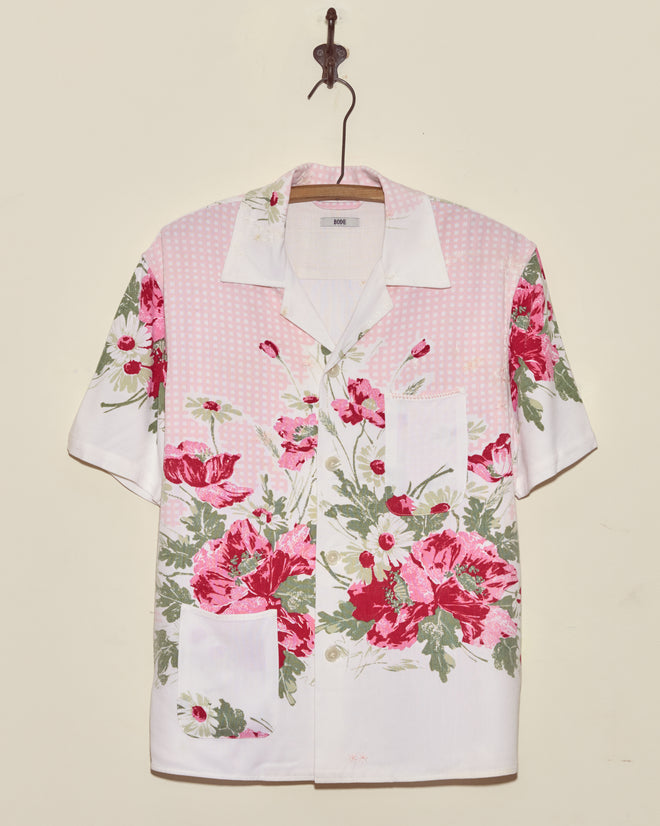 The Lovely Garden Short Sleeve Shirt (XS/S), featuring a pink and red floral print, is displayed on a wooden hanger against a beige wall.
