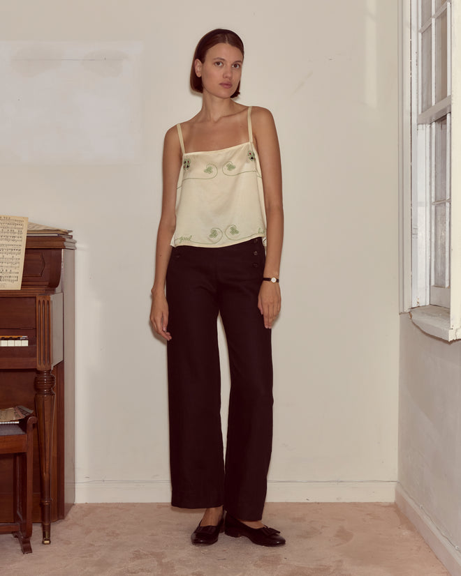 A woman wearing the Lucky Draw Tank—hand-embroidered silk with a subtle playing card motif—and black pants stands indoors by a piano and window, looking at the camera.