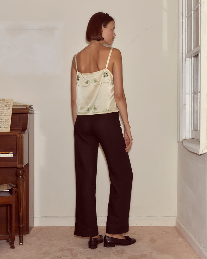 A woman with short hair stands indoors near a piano and window, facing away, wearing the Lucky Draw Tank featuring a subtle playing card motif and black pants.