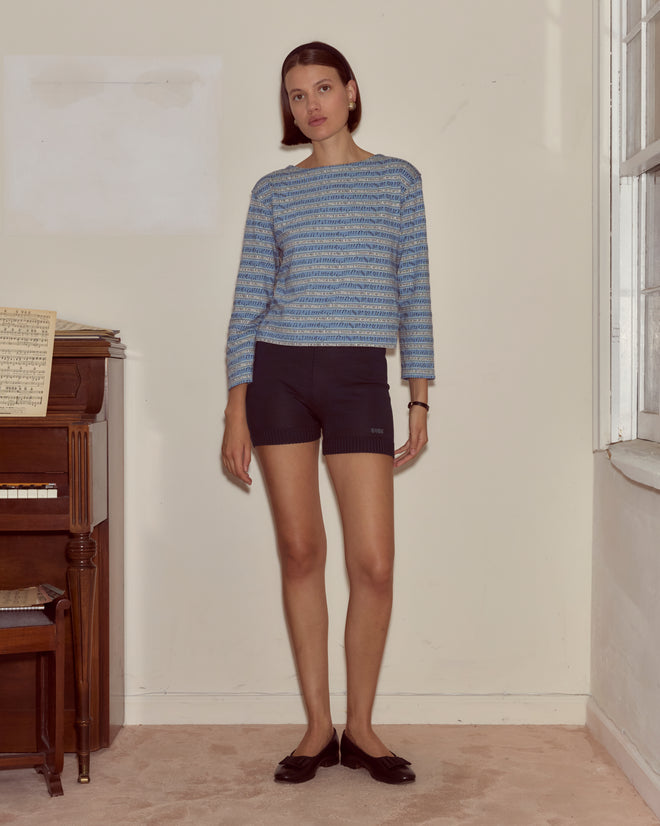 A woman in the Lyric Long Sleeve and shorts stands indoors by a piano with sheet music, channeling the expressive vibe of an artistic musical.