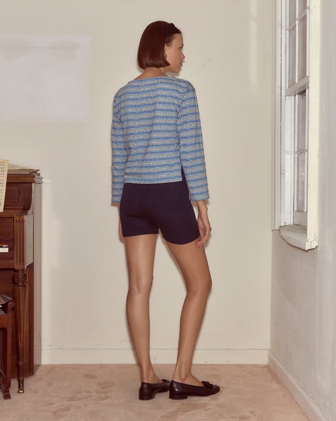 A woman stands in a corner facing the wall, wearing the Lyric Long Sleeve blue sweater with jacquard stripes, dark shorts, and black shoes.