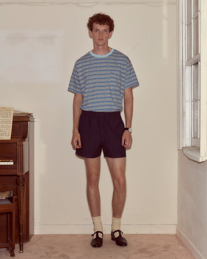 A young man in a Lyric Tee stands indoors near a piano and a large window.
