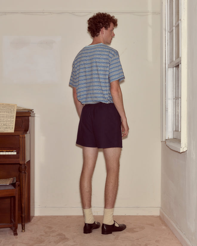 A man in a Lyric Tee and shorts stands facing the corner of a room near a piano and window.
