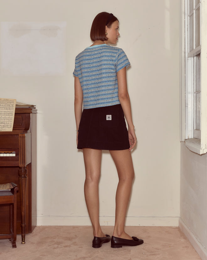 A woman in the Lyric Cropped Tee and a black skirt stands by a window, facing away from the camera.