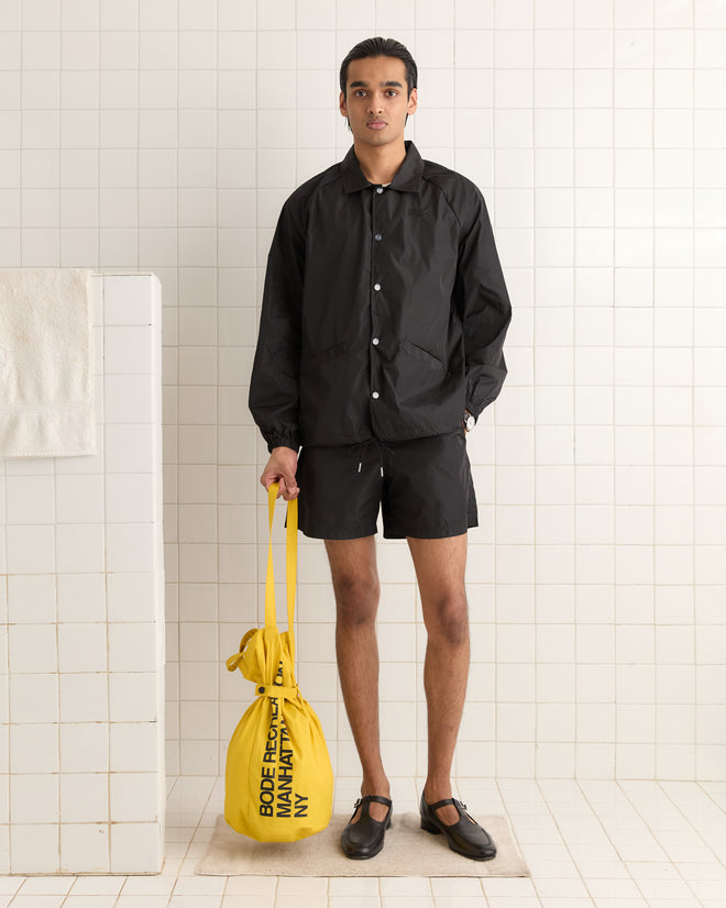 Dressed in a sleek black outfit, the individual stands in a tiled room holding a vibrant yellow Manhattan Island Bag.